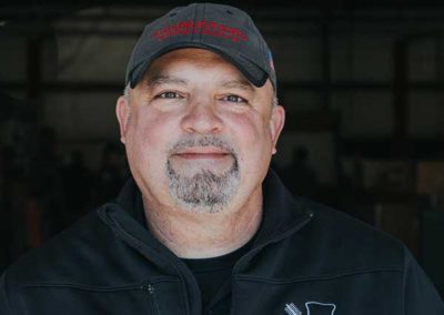 Shon Wilson - Logo hat and shirt with goatee and mustache. Grayish hair.
