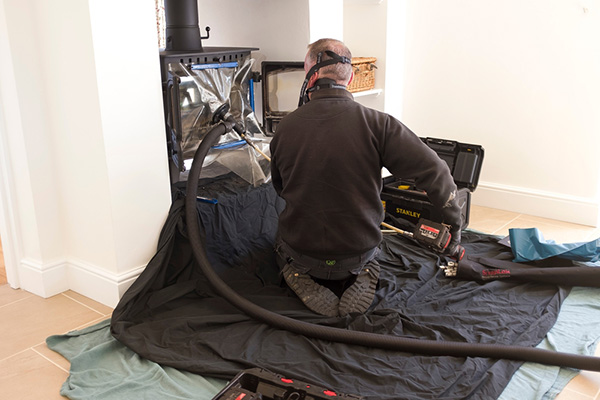 man kneeling on a black blanket with vaccum tubes connected to wood sttove cleaning it