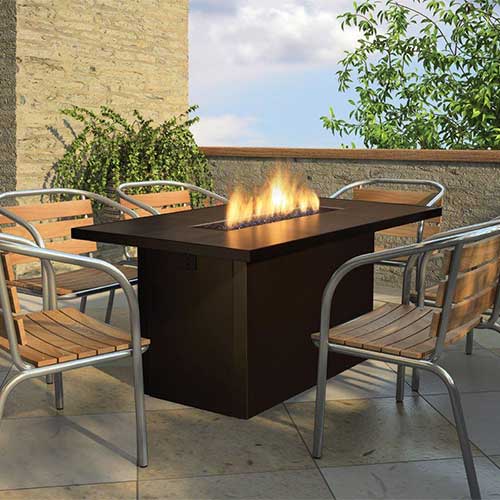 Five wooden chairs on a patio surrounding a black table with a rectangular open fire burning in it